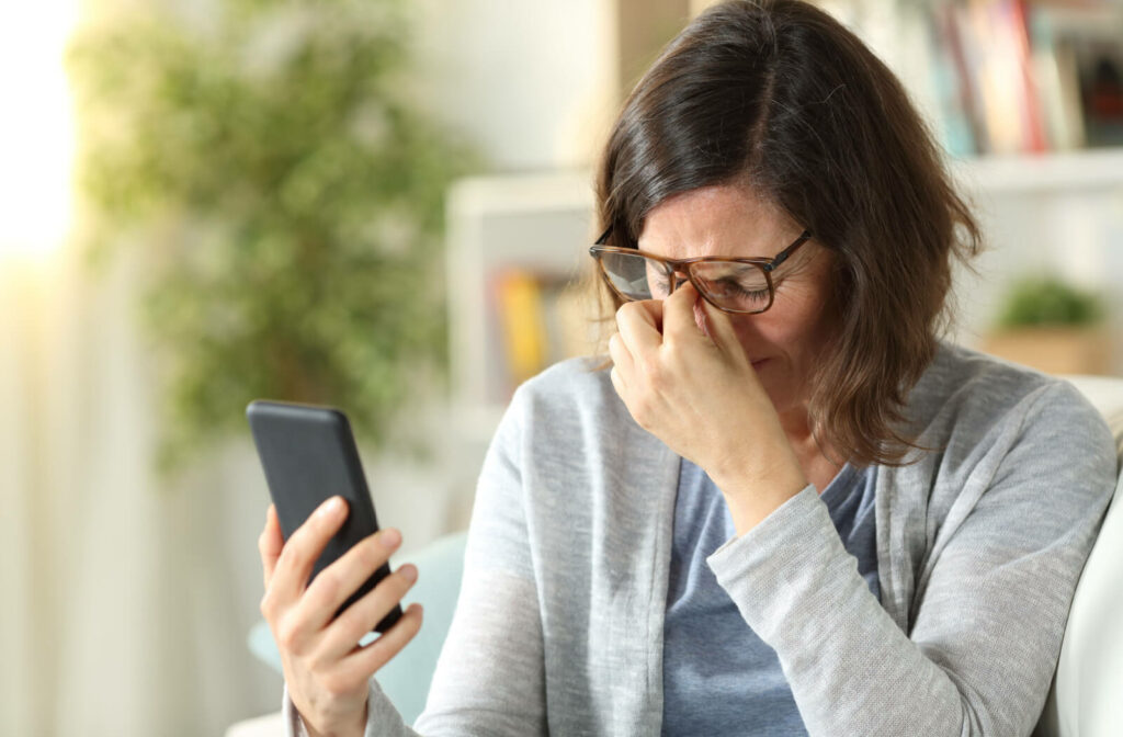 An adult woman with eyeglasses experiencing eye strain due to a long period of using her phone.