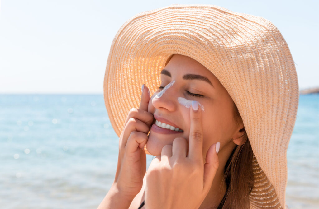 A woman wearing a hat at the beach putting sunscreen on her face.
