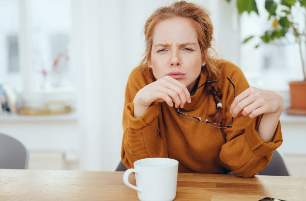 A woman in an orange sweater sitting at a table with a mug, holding her glasses in her hands and squinting to see.