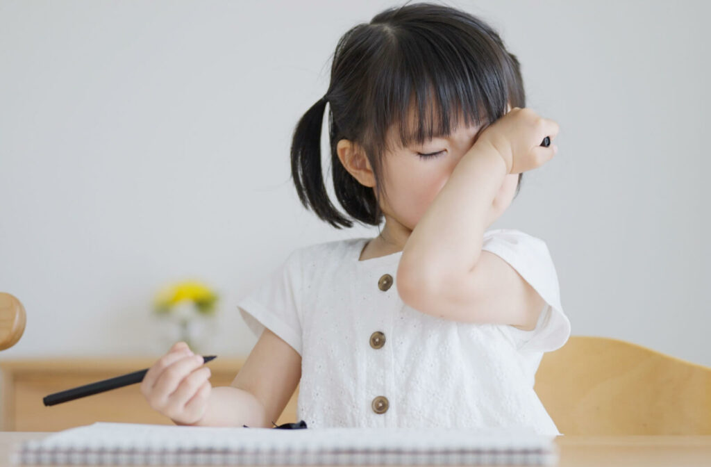 A young child sitting at a desk and rubbing her eyes while holding a pen in her right hand.