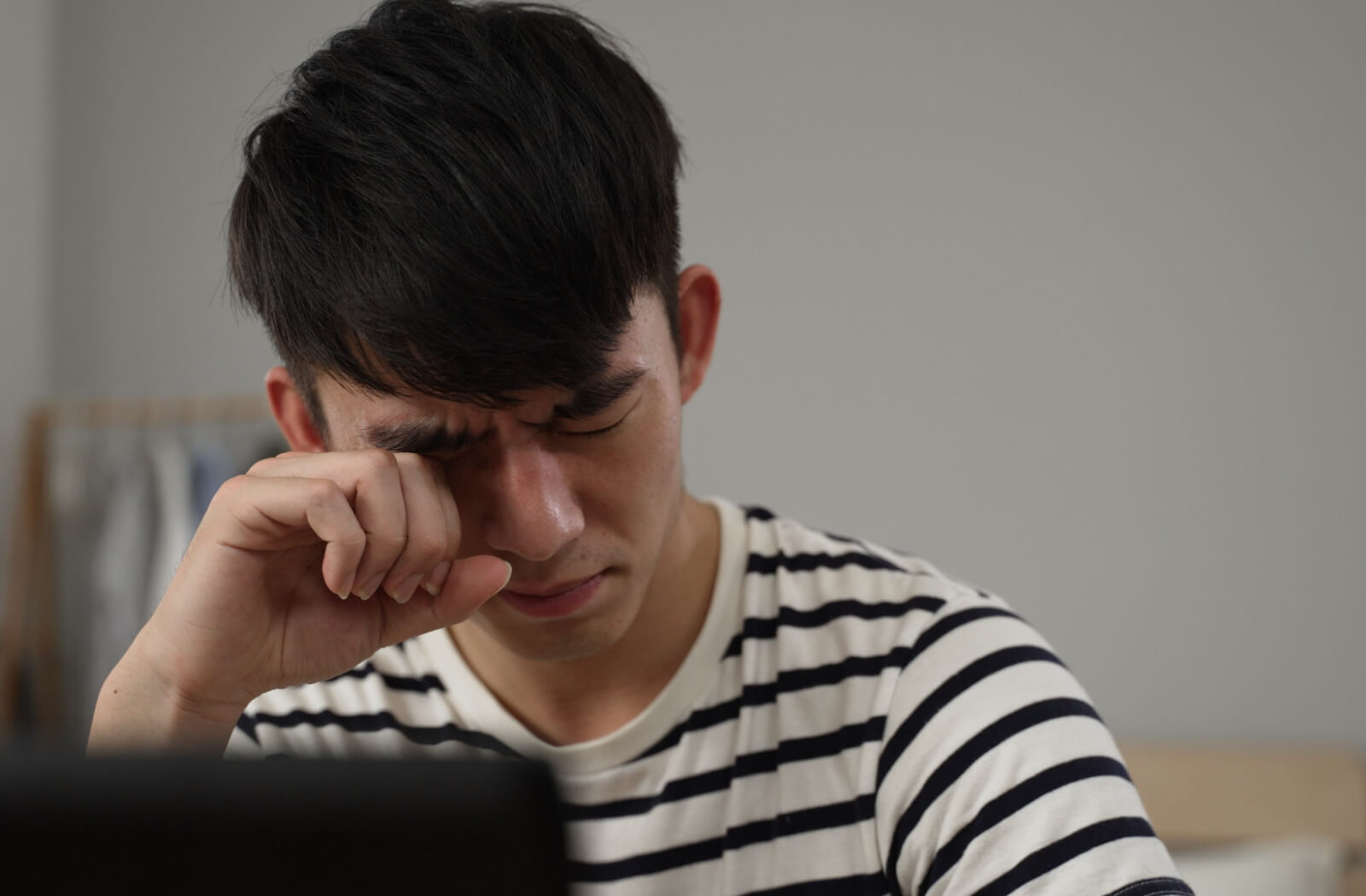 A young man sitting at a desk with his laptop as he rubs his eyes.