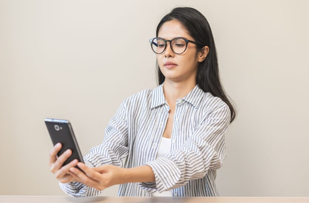 A woman wearing glasses holding her phone out as far as she can to be able to see clearly.
