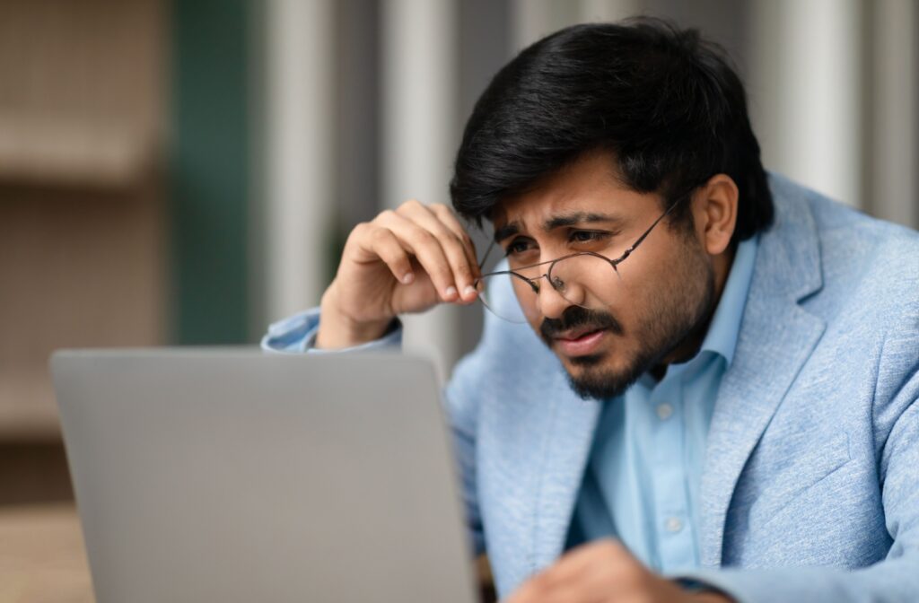 A young professional businessman squinting through eyeglasses, looking at a laptop. He appears to be suffering from nearsightedness.