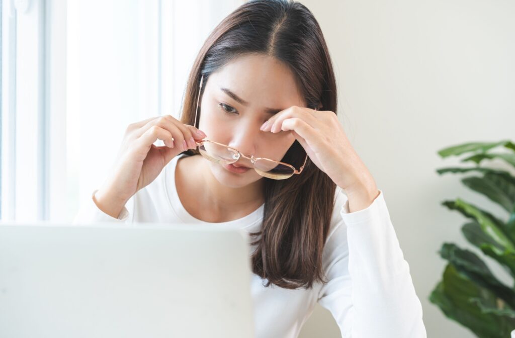 A woman sits in front of the computer holding her glasses in one hand and rubbing her eye with her other hand
