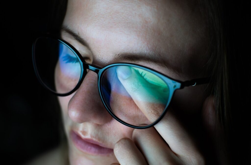 A person rubs their eyes while wearing glasses with a computer screen reflecting in the glasses lens