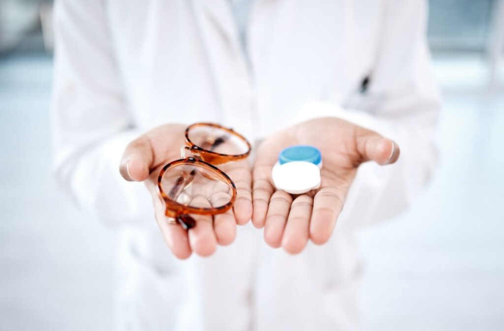 A person in a white coat holding a pair of eyeglasses in one hand and a contact lens case in the other.