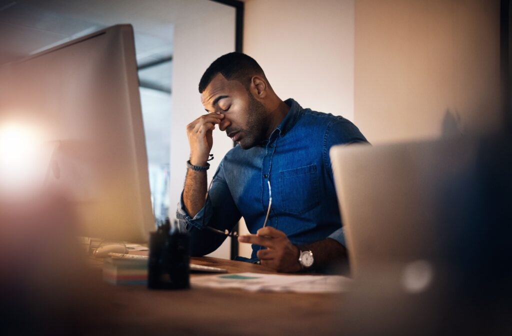 A person sits in front of a computer and rubs their dry eyes